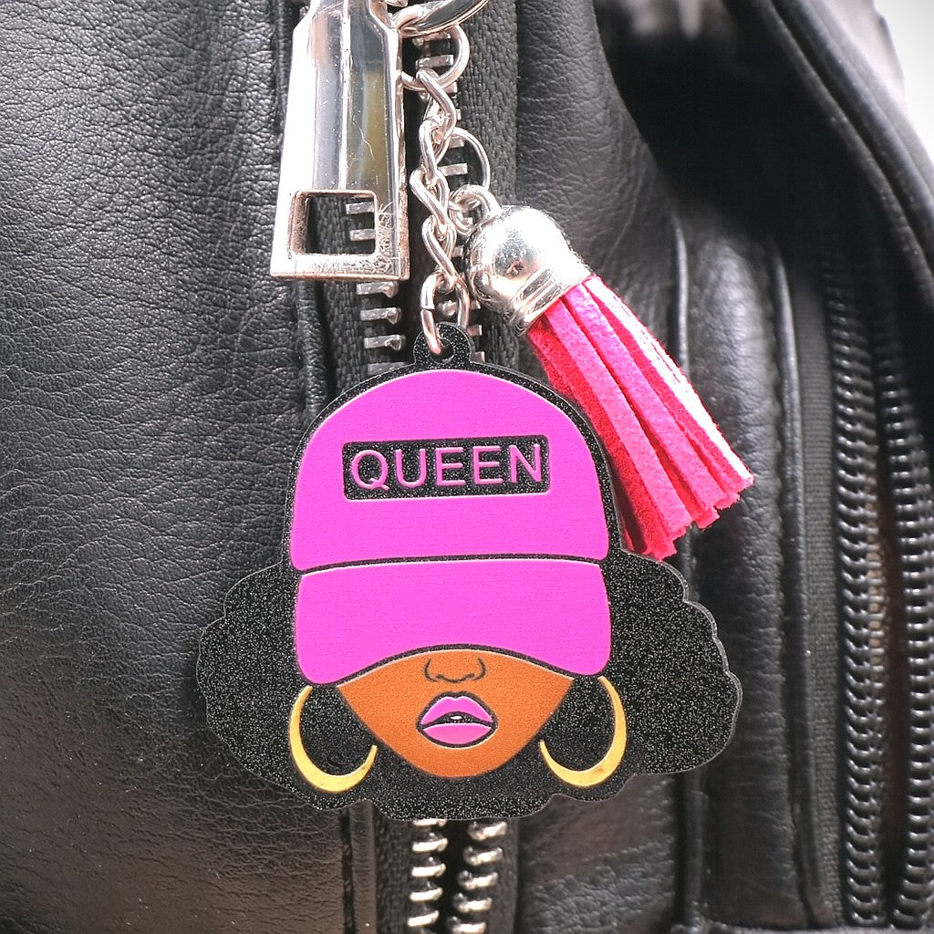 Keychain featuring a glittery illustration of a Black woman with curly hair, gold hoop earrings, and a hot pink cap labeled “QUEEN,” accented with a matching pink tassel and silver key ring on a black leather purse.