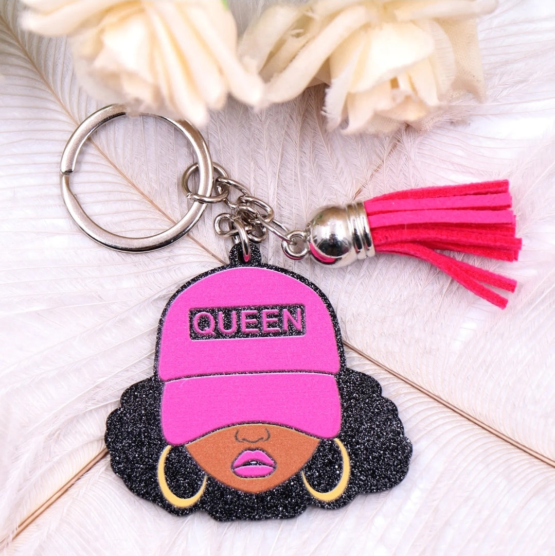 Keychain featuring a glittery illustration of a Black woman with curly hair, gold hoop earrings, and a hot pink cap labeled “QUEEN,” accented with a matching pink tassel and silver key ring on a light background.