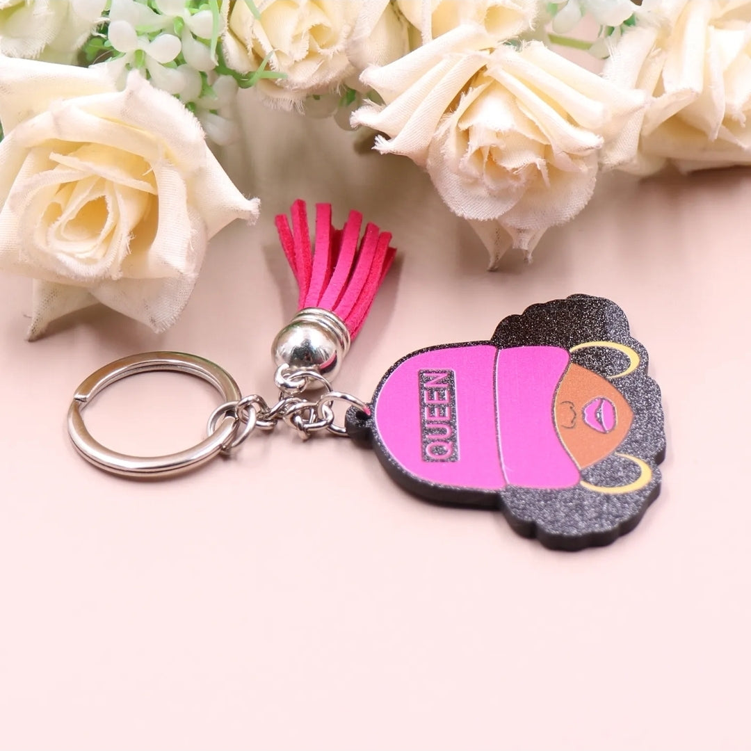 Side view of keychain featuring a glittery illustration of a Black woman with curly hair, gold hoop earrings, and a hot pink cap labeled “QUEEN,” accented with a matching pink tassel and silver key ring on a light background with flowers.
